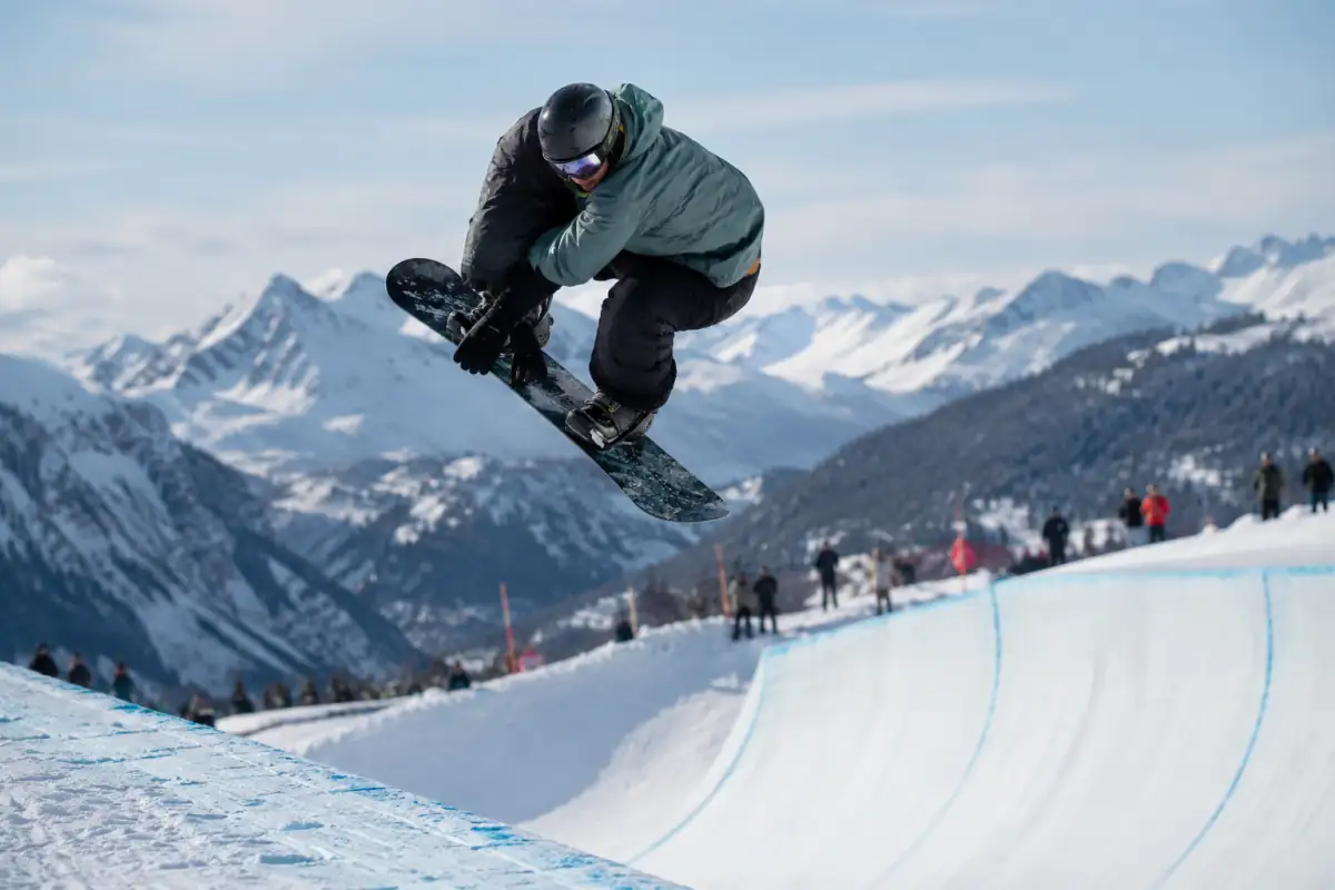 Snowboardeur en plein saut au-dessus d’une paroi enneigée dans un halfpipe de station alpine, équipé d’un casque et d’une planche freestyle.