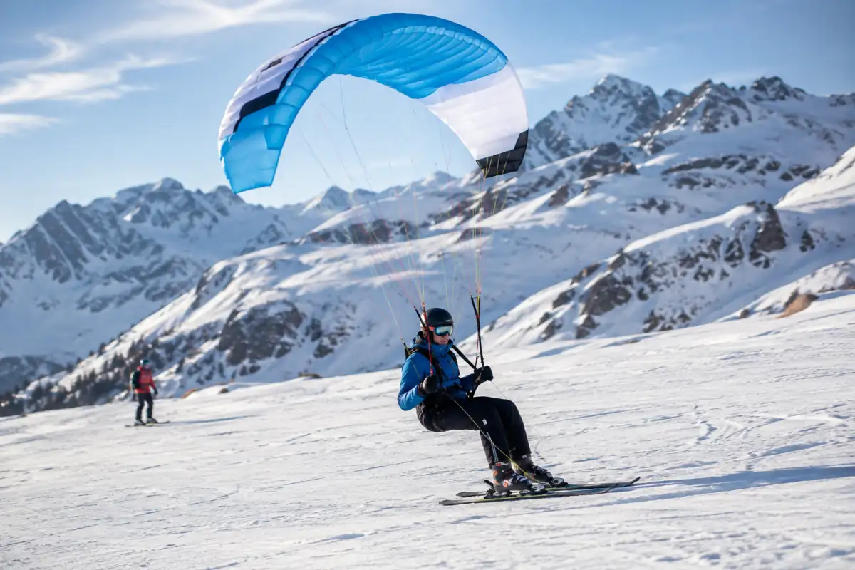 Un pratiquant de speed riding descend une pente enneigée à skis sous une petite aile de vol libre dans les Alpes.
