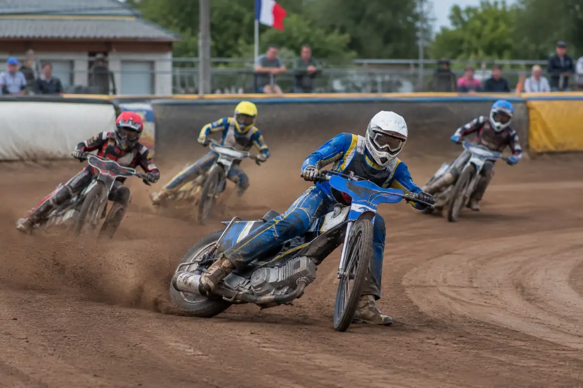 Un pilote de speedway moto négocie un virage en glisse sur une piste de terre, chaussure gauche équipée d’un patin.