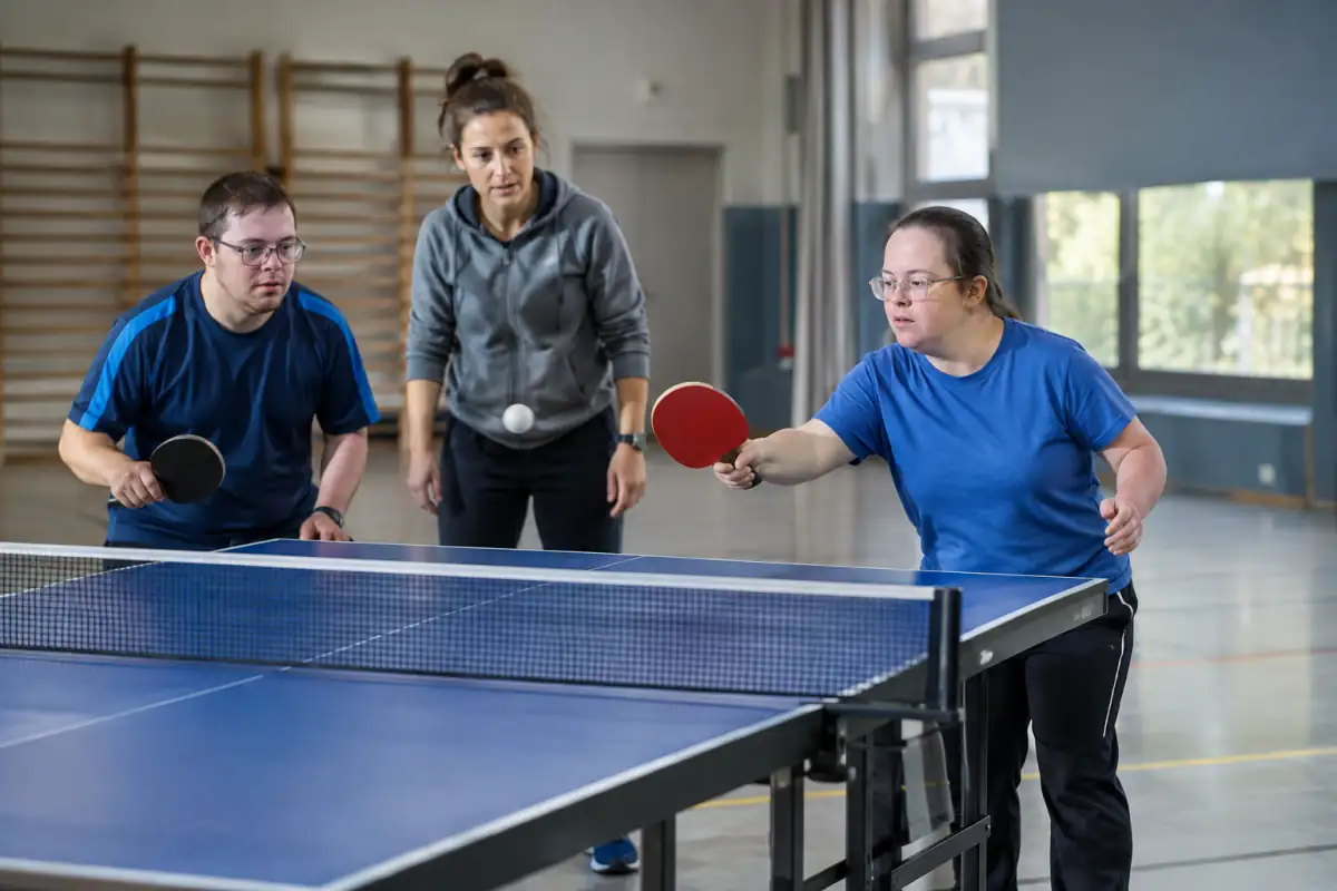 Deux adultes jouent un échange de tennis de table adapté dans une salle claire, avec un éducateur attentif à proximité.