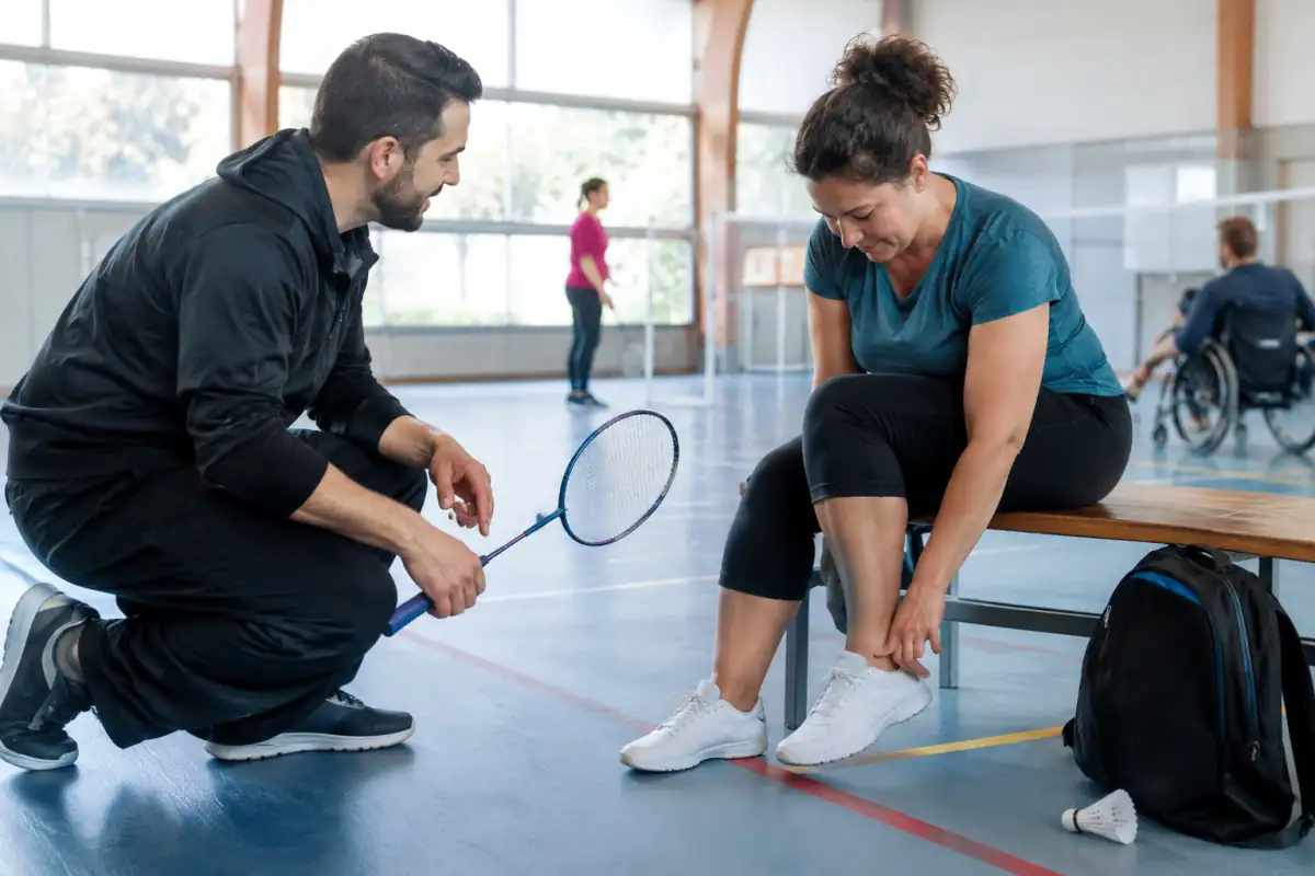 Dans un gymnase, une personne vérifie ses chaussures avant une séance de badminton adapté tandis qu’un encadrant lui prête une raquette.