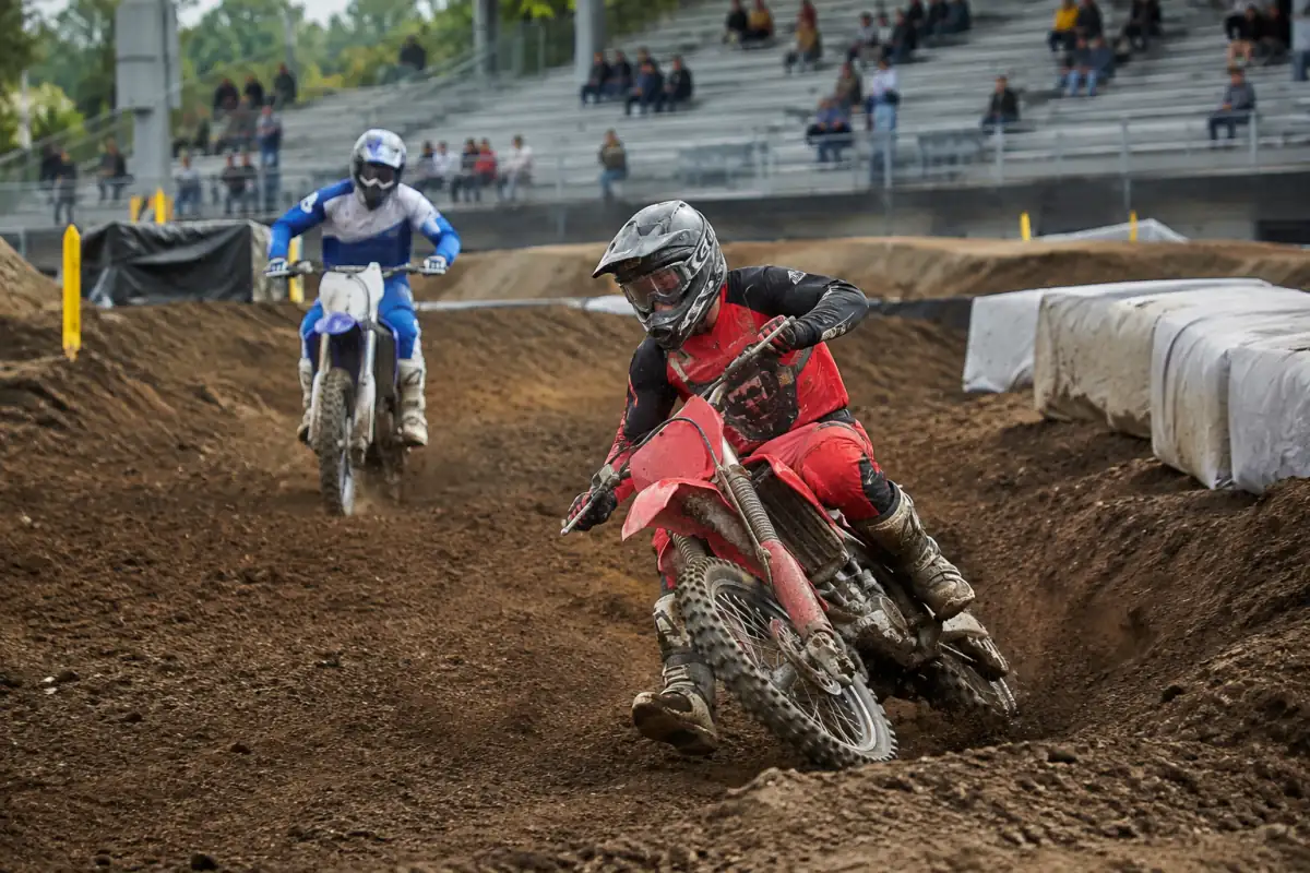 Deux pilotes en équipement de protection négocient un virage relevé sur un circuit compact de supercross aménagé dans un stade.