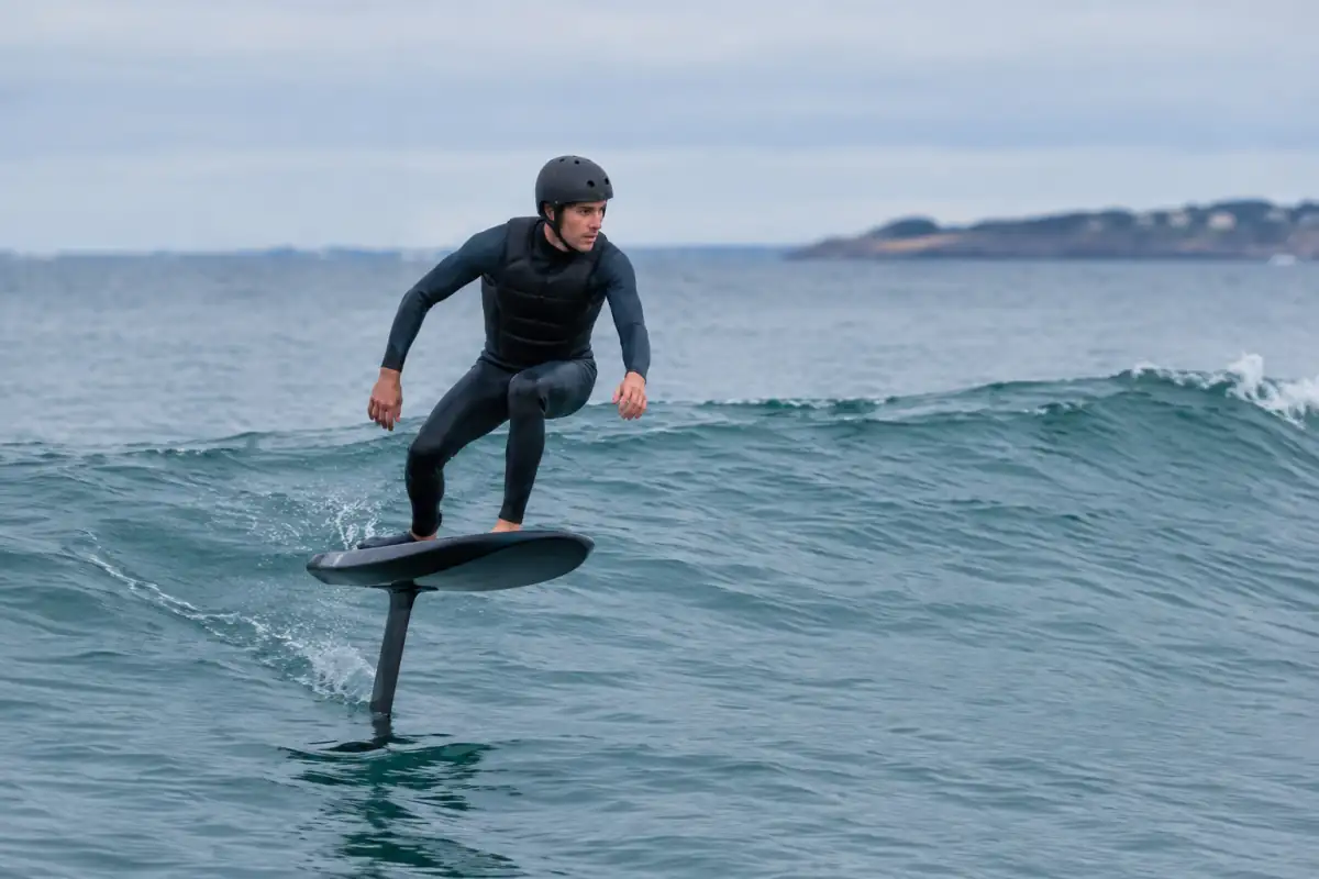 Un pratiquant en combinaison et casque glisse au-dessus d’une vague sur une planche de surf foil équipée d’un mât.