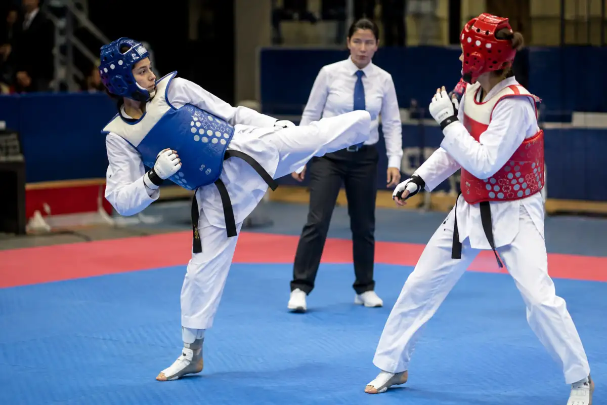 Deux combattants en protections WT exécutent un coup de pied contrôlé au plastron dans une aire de compétition de taekwondo.