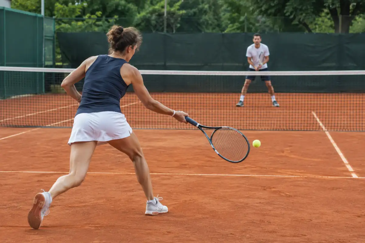 Échange de tennis en simple sur terre battue : une joueuse frappe un coup droit pendant que son adversaire se prépare.