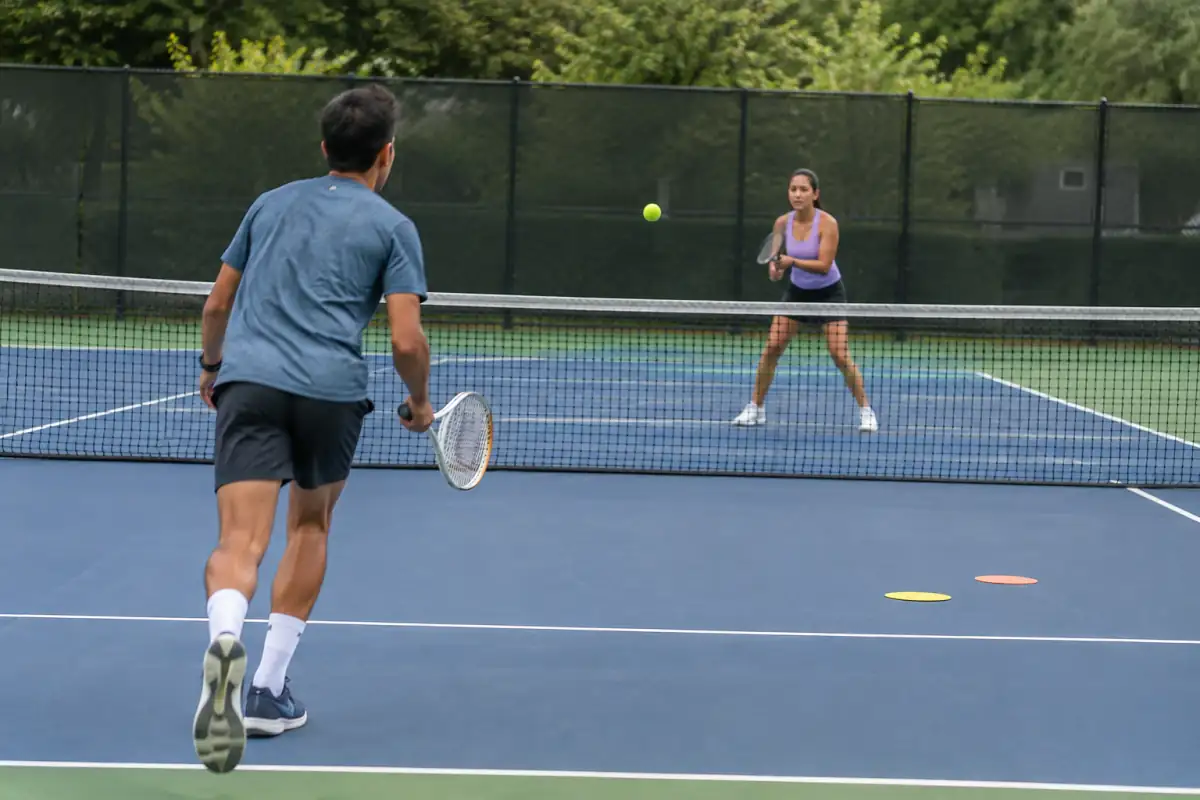 Deux joueurs débutants échangent sur un court de tennis, l’un se replace après sa frappe tandis que l’autre prépare son retour.
