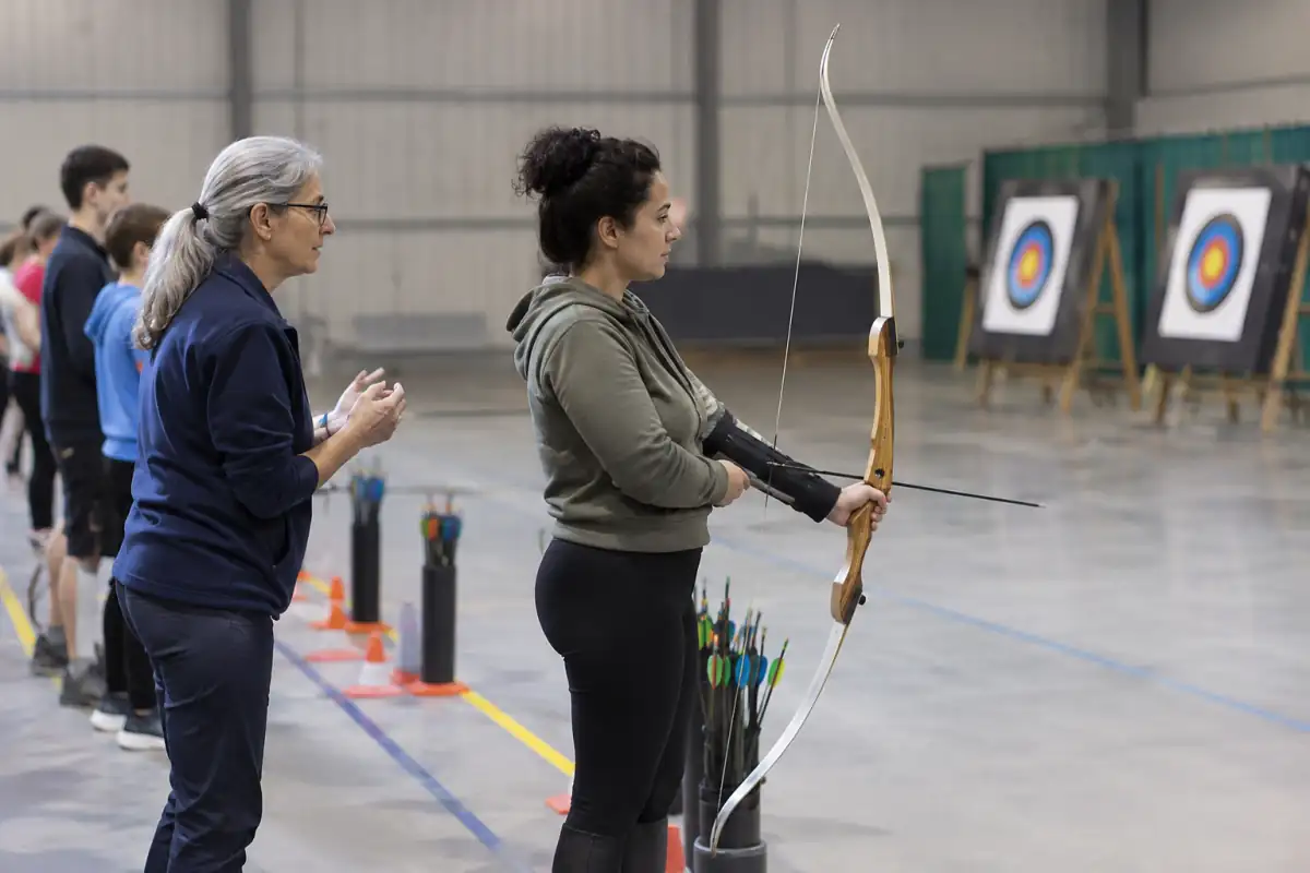 Un débutant écoute un entraîneur au pas de tir d’un club, arc prêté en main et zone d’attente visible derrière la ligne.