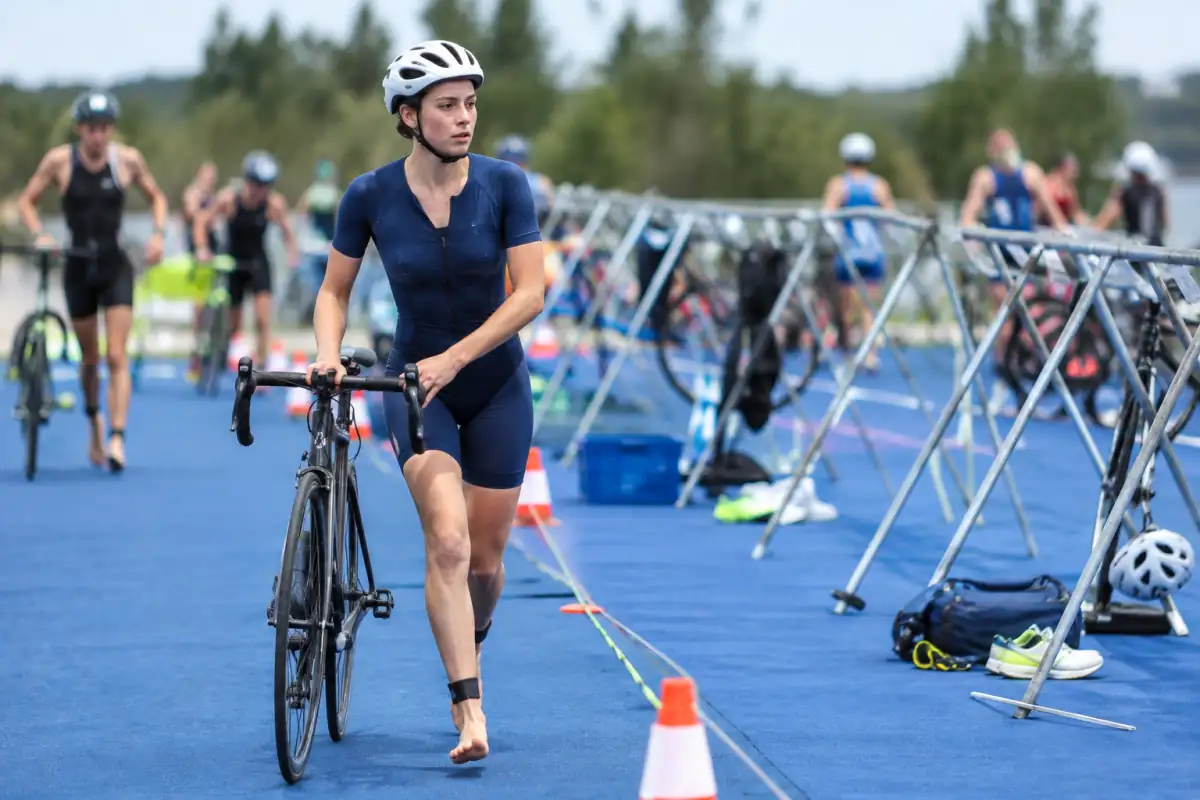 Triathlète casqué poussant son vélo à pied dans l’aire de transition, avant la ligne de montée et avec son matériel rangé.