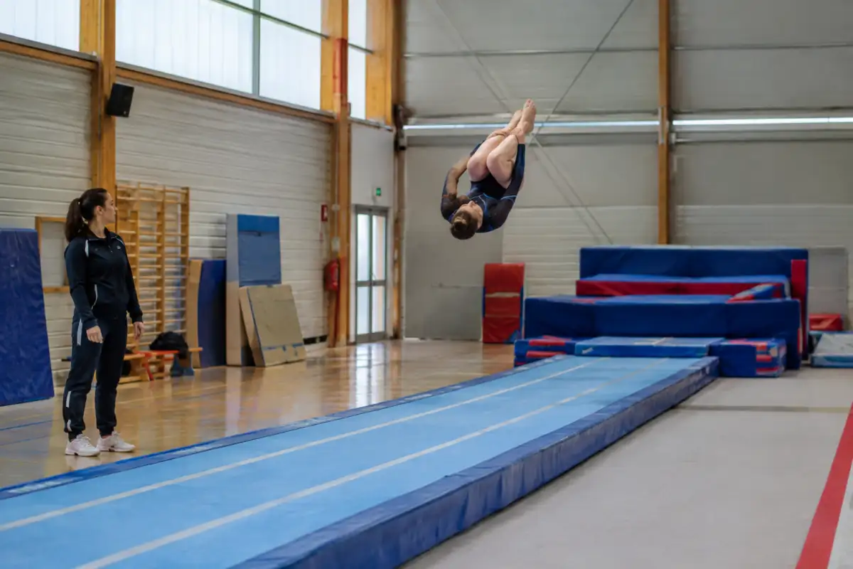 Une gymnaste enchaîne un flip et un salto sur une piste de tumbling dynamique, sous la surveillance d’un entraîneur en salle.