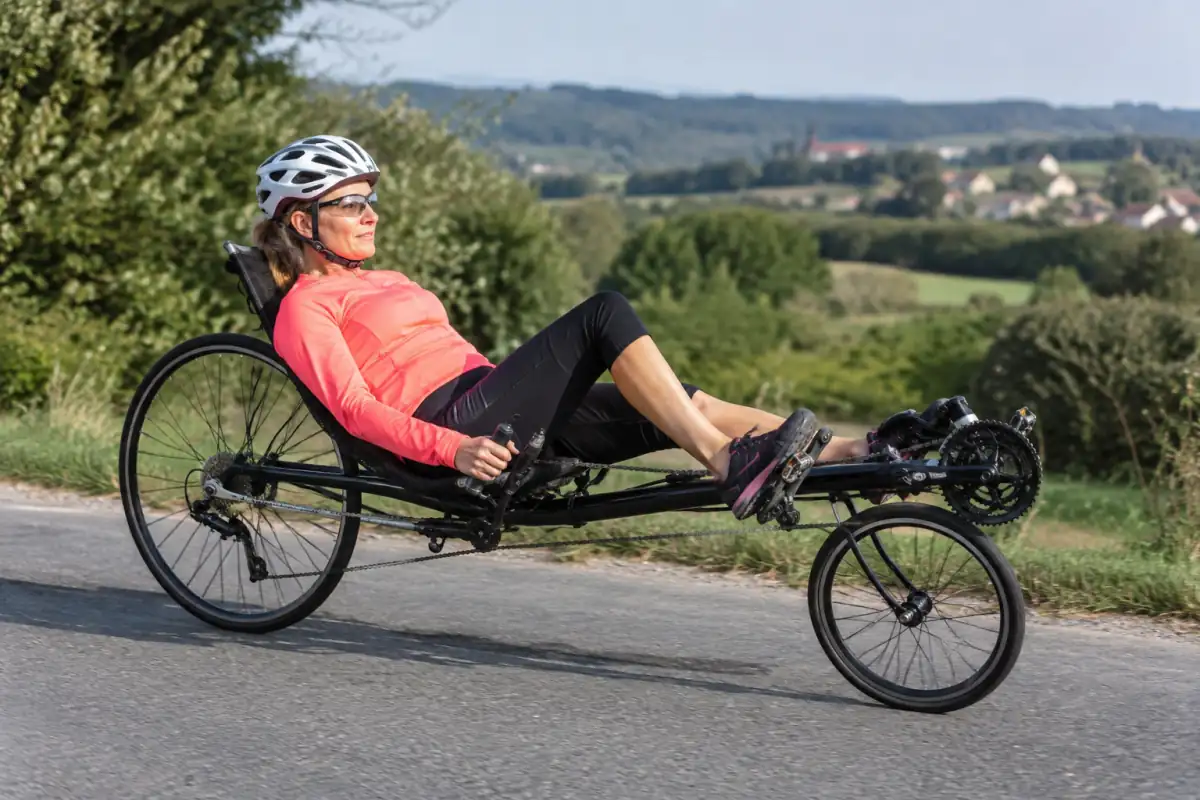 Un cycliste en vélo couché pédale sur une route de campagne, le dos soutenu par le siège et les jambes tendues vers l’avant.