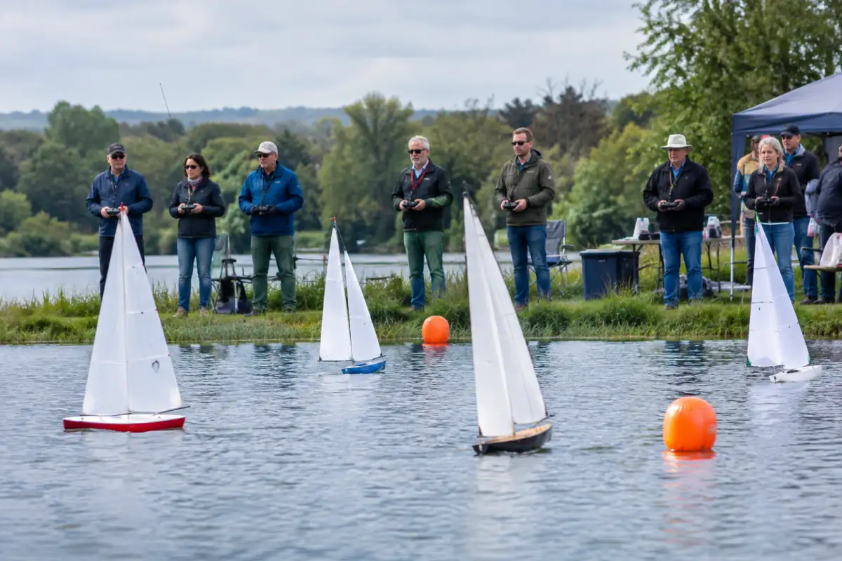 Des skippers pilotent leurs voiliers de voile radiocommandée depuis la berge pendant une régate sur un plan d’eau.