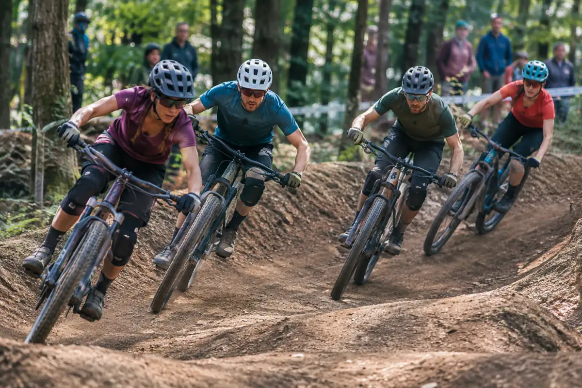 Quatre pilotes de VTT cross-country eliminator négocient un virage serré sur un parcours en terre, casqués et protégés.