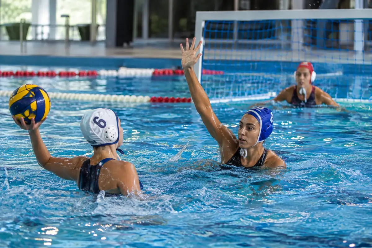 Un joueur de water-polo prépare une passe près de la ligne des 5 mètres, face à un défenseur et au but.
