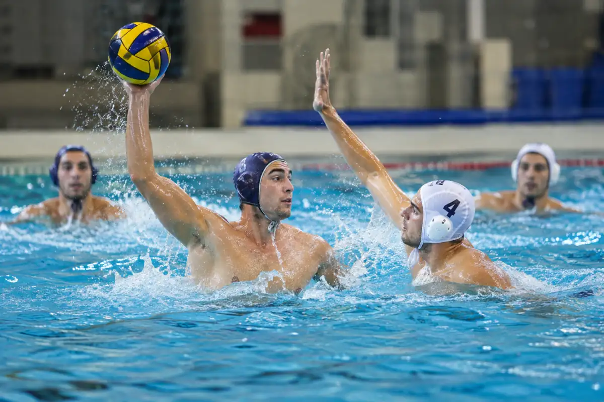 Joueur de water-polo en appui haut, réceptionnant le ballon d’une main sous la pression d’un défenseur avant une passe.