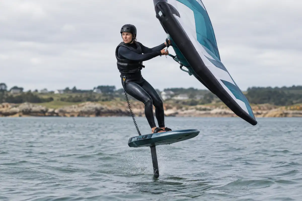 Un pratiquant de wingfoil vole au-dessus de l’eau avec une aile tenue à la main, sur un plan d’eau côtier breton.