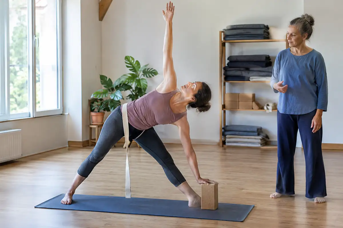 Une élève tient une posture debout de yoga Iyengar, la main posée sur une brique dans un studio encadré par un professeur.