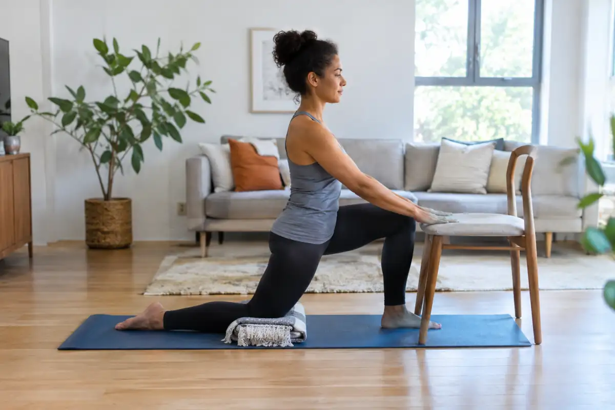 Débutante pratiquant une fente basse de yoga chez elle, genou posé sur une couverture et mains en appui sur une chaise.